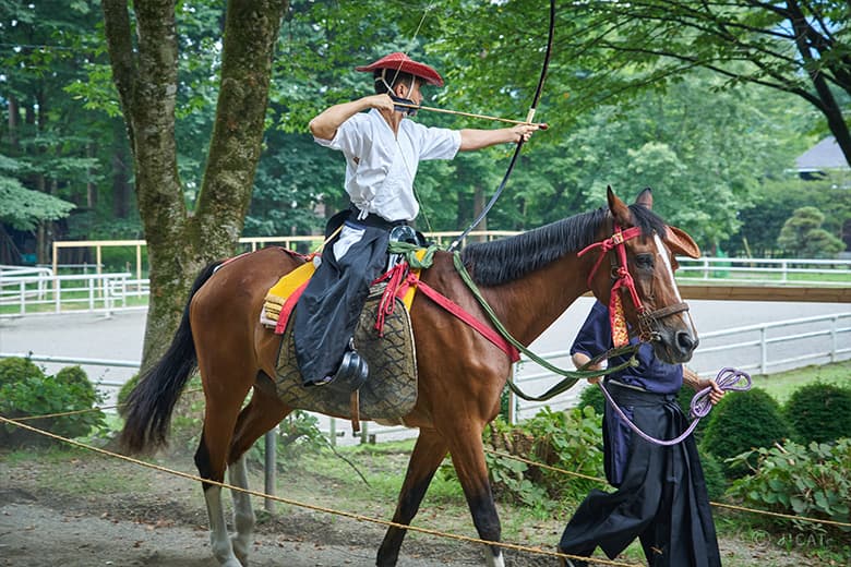 A yabusame trainee practicing horseback archery, drawing a bow while riding a horse under the guidance of an instructor in a countryside setting