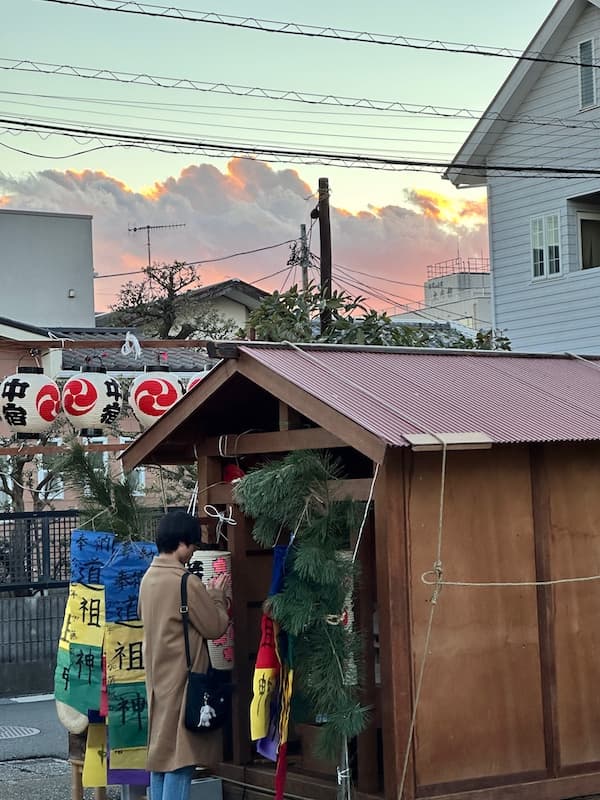 A person praying at a wooden okariya shrine during the Sagicho Festival in Oiso, with colorful decorations and lanterns.
