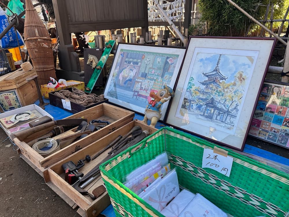 Vintage goods and artwork for sale at a flea market in Tokyo, including framed drawings, wooden tools, and household items in crates.