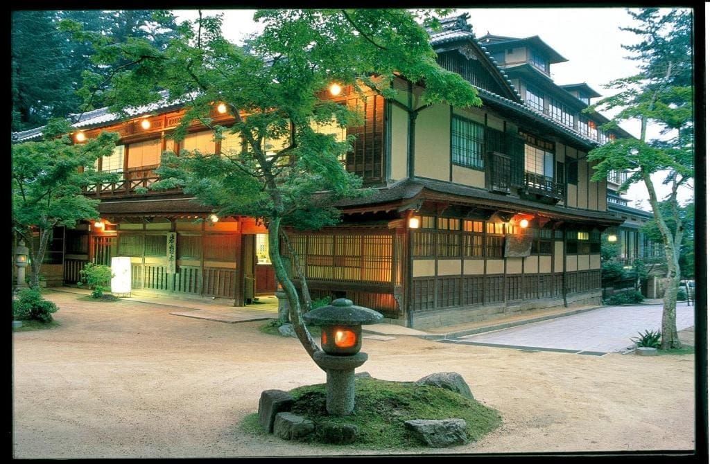 Traditional wooden exterior of Iwaso ryokan with warm lighting, surrounded by trees and a stone lantern in the front garden.