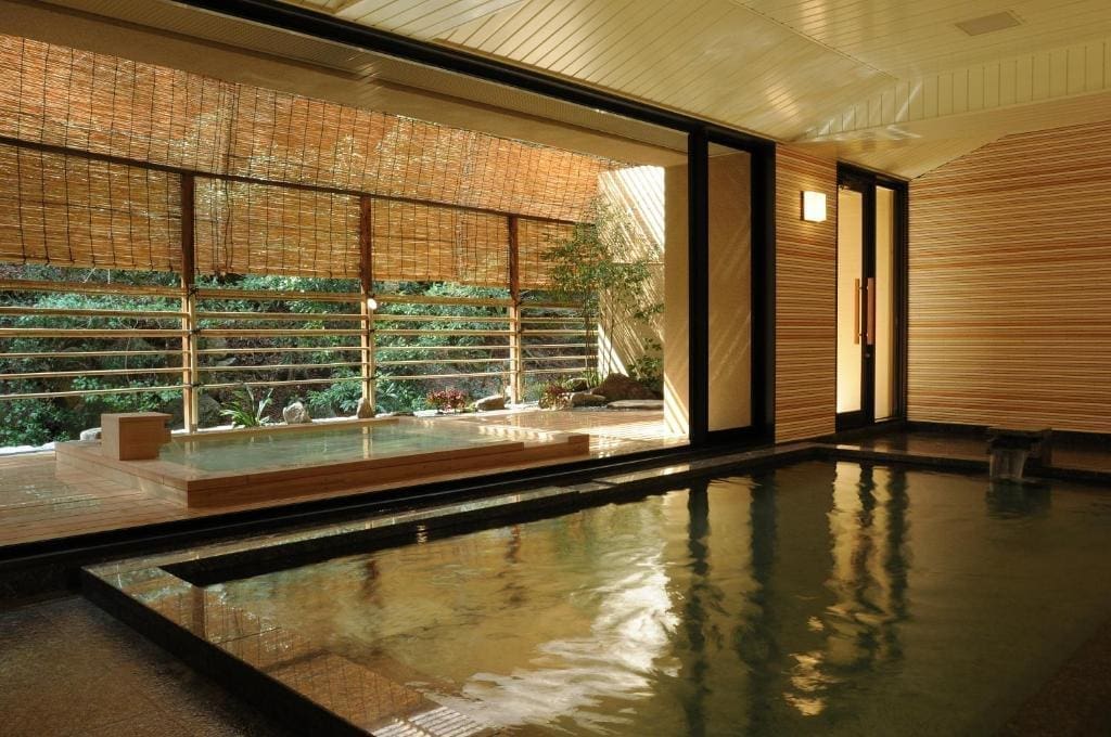 Indoor and outdoor bath area at Iwaso with open windows looking out to a garden, featuring bamboo blinds and tiled floors.