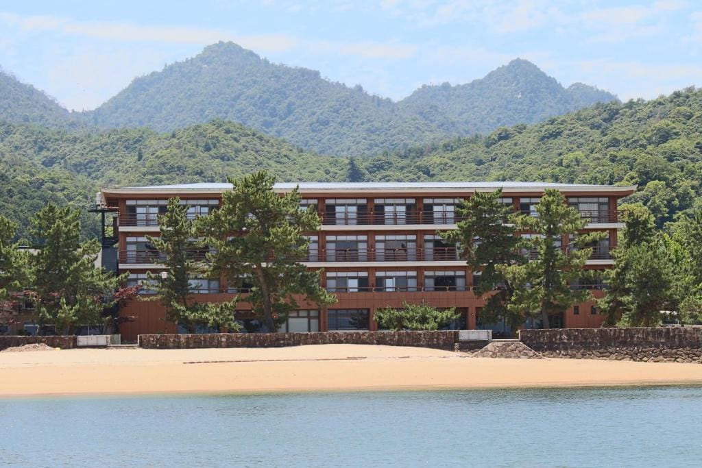 Exterior view of Miyajima Seaside Hotel with balconies facing the water, located along a quiet beach with forested hills behind it.