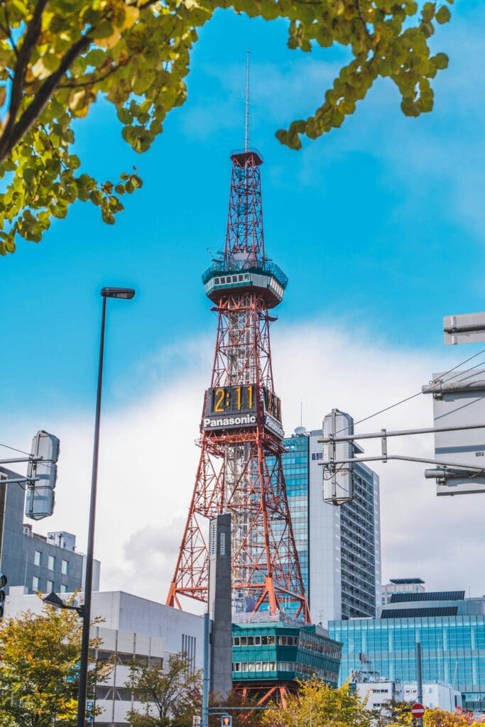Red Sapporo TV Tower with Panasonic digital clock rising above city buildings and autumn trees under a blue sky