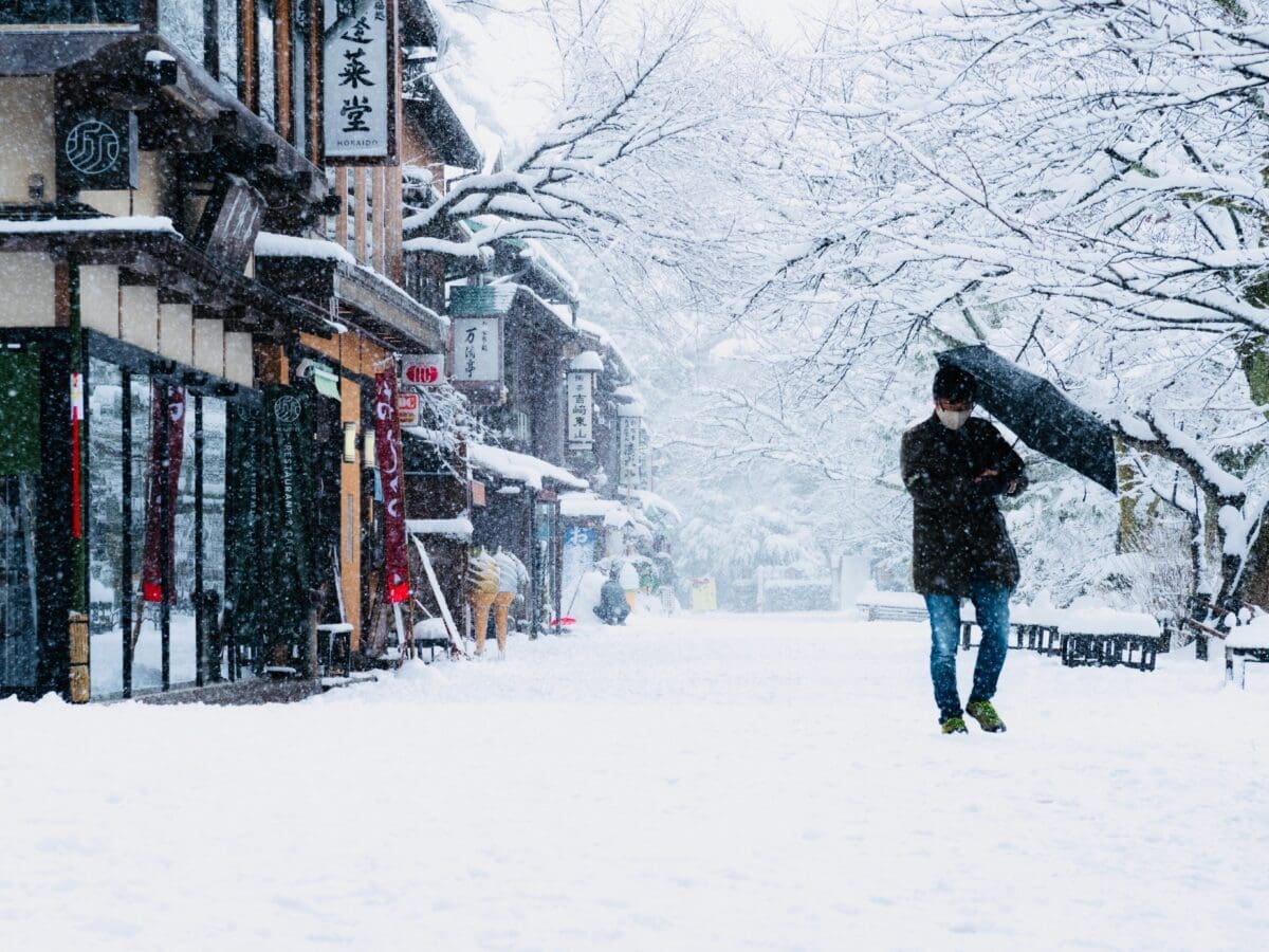 Person holding umbrella walking down snow-covered street with Japanese storefronts in Kanazawa