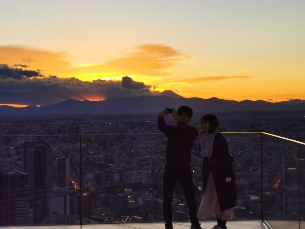 Couple taking a selfie at Shibuya Sky observation deck during sunset with Tokyo cityscape and Mount Fuji silhouette in the background