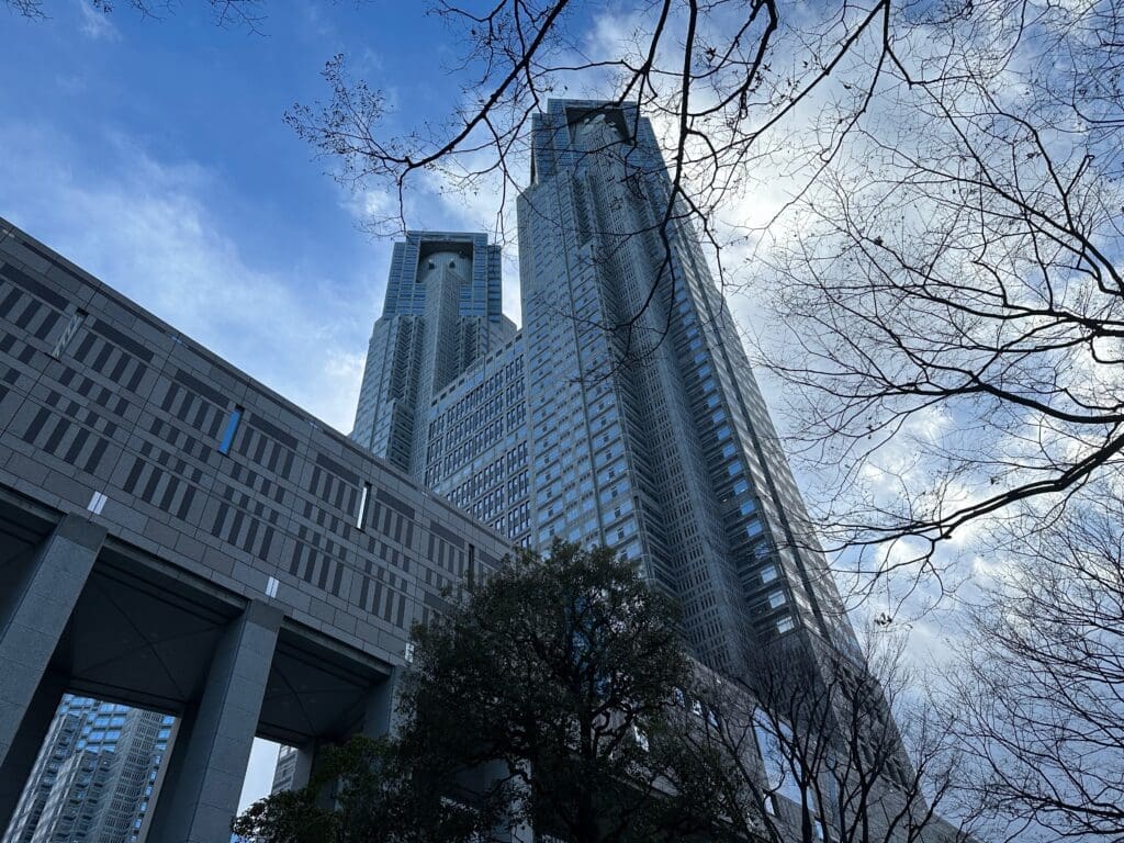 Low-angle view of Tokyo Metropolitan Government Building twin towers framed by bare tree branches under a cloudy blue sky