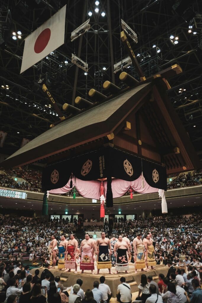 Sumo wrestlers lined up in a dohyo under a suspended Shinto shrine roof at Ryogoku Kokugikan in Tokyo