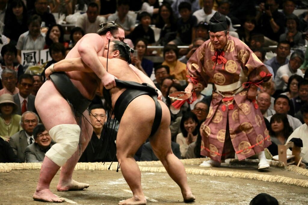 Two sumo wrestlers grappling during a match in a Japanese sumo arena with referee and audience