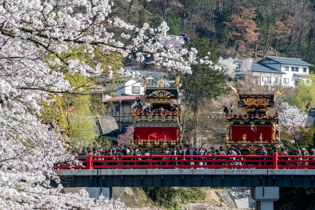 People and ornate festival floats on a red bridge in Takayama surrounded by cherry blossoms