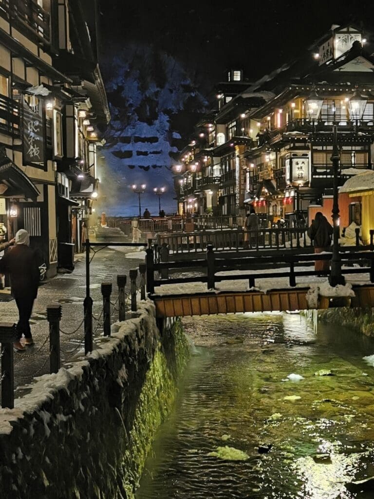Ginzan Onsen at night in snow, vertical view of lit ryokan inns and a bridge along the river in Yamagata, Japan