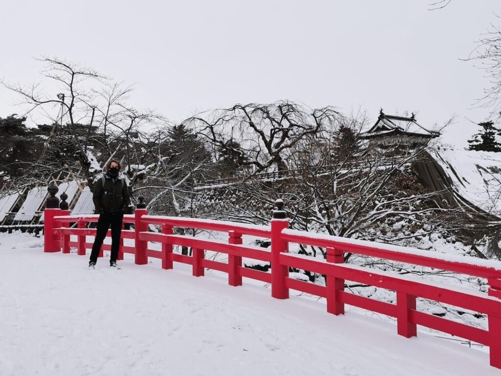 Person on a red bridge in winter at Hirosaki Castle, Aomori, Japan, with snow-covered trees and a castle turret