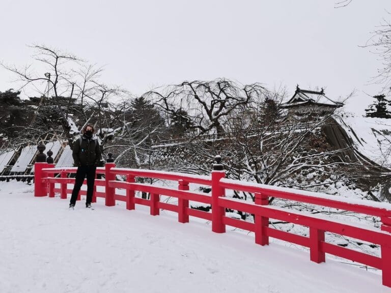 Person on a red bridge in winter at Hirosaki Castle, Aomori, Japan, with snow-covered trees and a castle turret