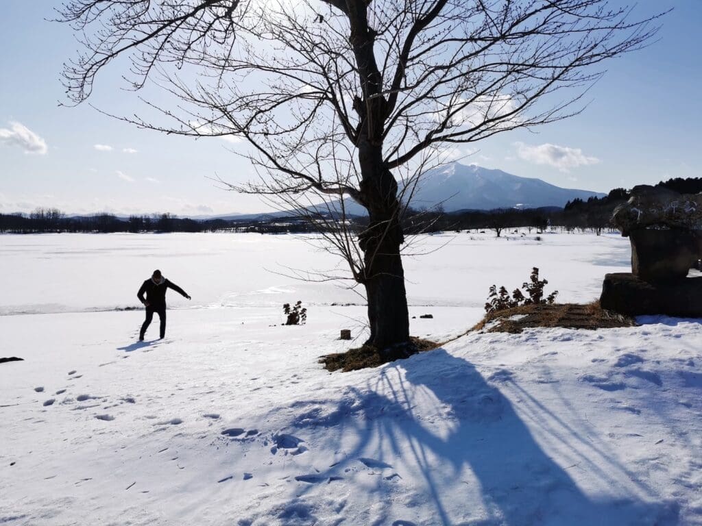 Mawarizeki Otame Pond frozen in winter, a person walking on snow beside a bare tree with mountains in the background