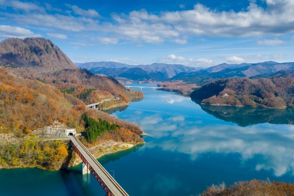Aerial view of a blue mountain reservoir with autumn forests, a railway bridge entering a tunnel in the foreground, and a highway bridge in the distance in Semboku, Akita, Japan