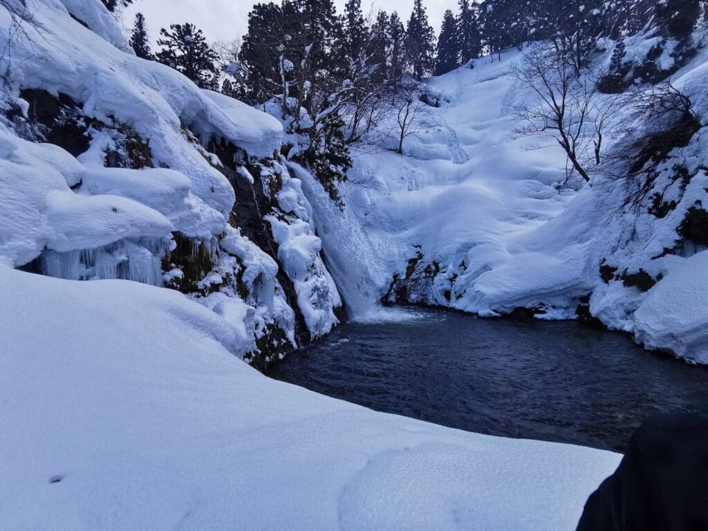 Shirogane Falls in winter at Ginzan Onsen, Yamagata, with snow-covered cliffs, icicles, and a partially frozen cascade flowing into a dark pool